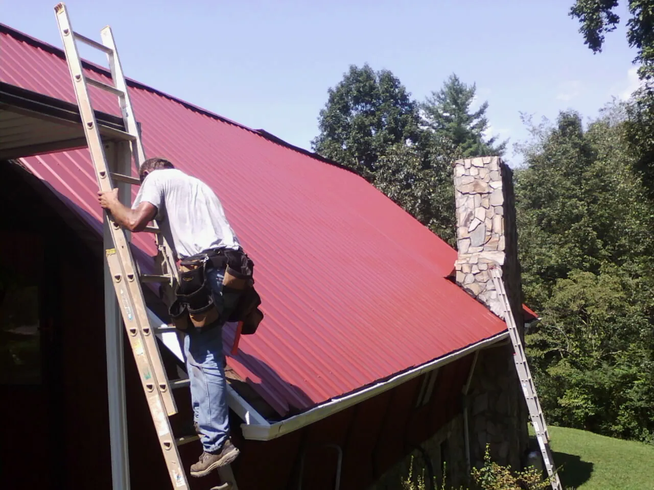 Roofer on a ladder beside a red metal roof and stone chimney — Soffit & Fascia Repair by Roofing Brothers in Allison